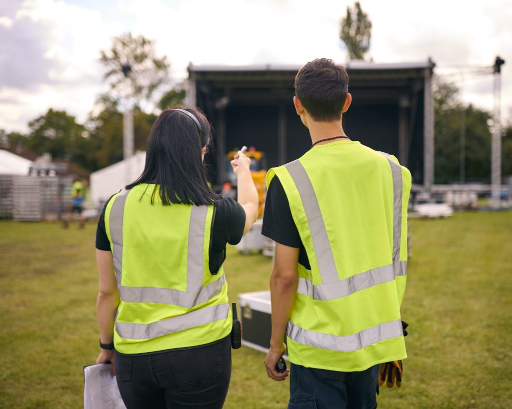 Rear View Of Production Team With Headsets Setting Up Outdoor Stage For Music Festival Or Concert